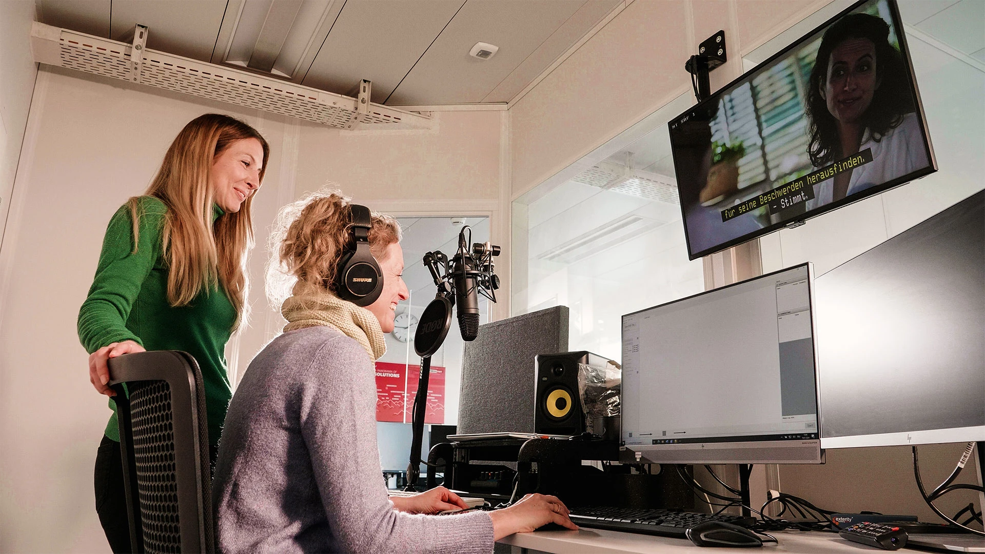 Eine Frau sitzt und eine Frau steht in einem Audioaufnahmeraum vor mehreren Bildschirmen. Beide Personen sind glücklich und schauen auf die Monitore. Die sitzende Person spricht in ein Mikrofon und audiodeskribiert einen Videobeitrag, der auf einem grossen Bildschirm läuft.